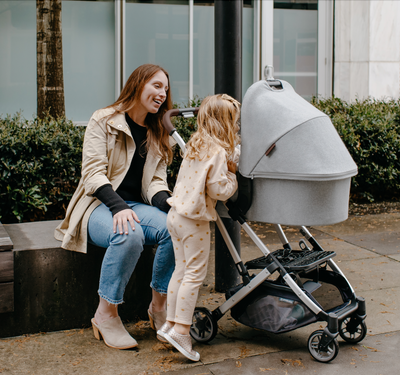 Mom and child looking into a bassinet attached to a stroller while sitting on an outdoor bench.